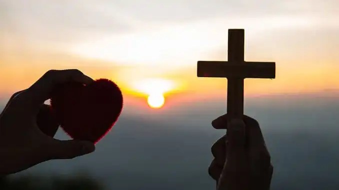 A breathtaking sunset with warm, glowing colors as a person holds up a heart-shaped cross in the foreground, symbolizing love, faith, and hope.
