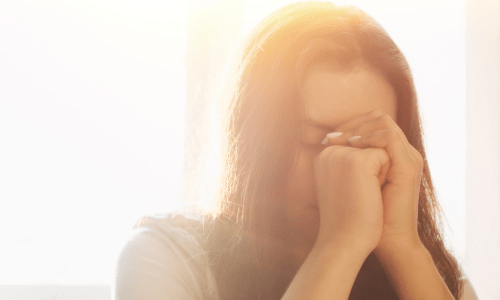 Woman with eyes closed and hands clasped in prayer, sitting quietly in soft natural light during a moment of gratitude and reflection with God.